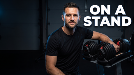 Chris Whelan resting his arm on adjustable dumbbells sitting on a waist-height stand, with the caption On a stand